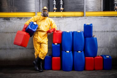 Tehlikeli madde giysisi ve gaz maskesi giymiş profesyonel bir işçinin portresi kimya fabrikasında duruyor ve kutuları tutuyor..