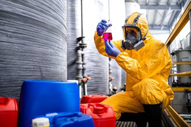 Chemical factory worker wearing protection suit and gas mask testing quality of chemicals. In background storage tanks for acid and industrial interior.