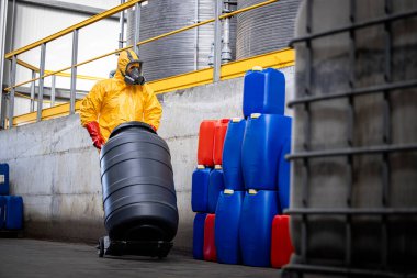 Modern kimya fabrikasında tehlikeli kimyasalları taşırken koruyucu giysi ve gaz maskesi takan işçiler. Arka planda asitli büyük tanklar.