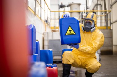 Factory worker in protective equipment and gas mask holding plastic can with toxic chemicals and biohazard waste inside recycling plant.