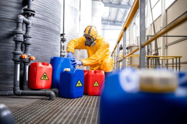 Factory worker in hazmat suit and gas mask filling plastic container with chemical and hazardous waste. Chemicals factory interior with acid storage tanks.