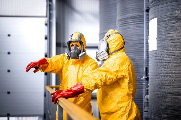 stock image Factory worker standing by large metal storage tanks with acids wearing yellow protection suit, gas mask and gloves explaining trainee process of chemicals production inside the plant.