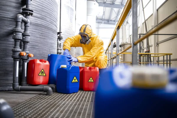 Factory worker in hazmat suit and gas mask filling plastic container with chemical and hazardous waste. Chemicals factory interior with acid storage tanks.