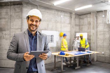 İnşaat mühendisleri arka planda plan yaparken elinde dijital tablet bilgisayarla deneyimli bir mimarın portresi.