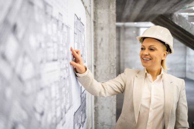 İş elbisesi ve güvenlik kaskı giymiş güzel kadın mühendis inşaat alanındaki proje planını analiz ediyor..