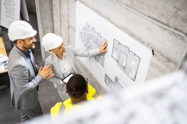 Bir grup mimar ve inşaat işçisi toplantı yapıyor, planları tartışıyorlar. İnşaat alanı içi.