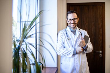 Hastanenin penceresinde duran beyaz üniformalı ve steteskoplu deneyimli bir doktorun portresi..