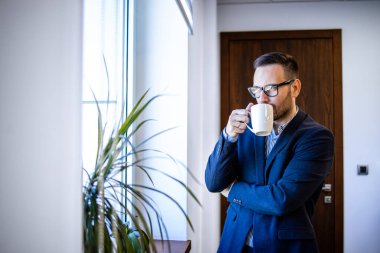 Ofisinde duran, kahve içen ve pencereden dışarı bakan başarılı yönetici ya da yönetmenin portresi..