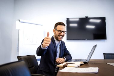 Pozitif ve neşeli iş adamı şirket içinde dizüstü bilgisayarı üzerinde çalışırken başparmağını kaldırıyor..