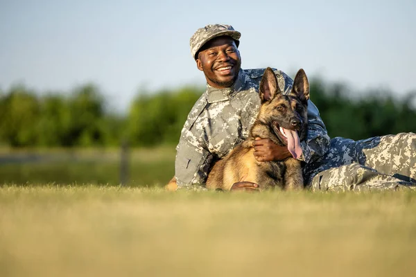 Soldier in military uniform and trained dog lying on grass playing and hugging.