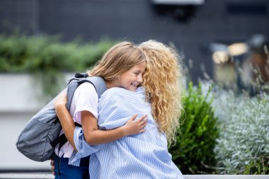 Anne ve kızı okula gitmeden önce kucaklaşıp öpüşüyorlar. Ebeveynler ve çocuklar arasında sevgi.