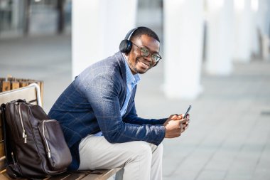Zarif takım elbiseli yakışıklı adam müzik dinliyor ve tren istasyonunda akıllı telefon kullanıyor..