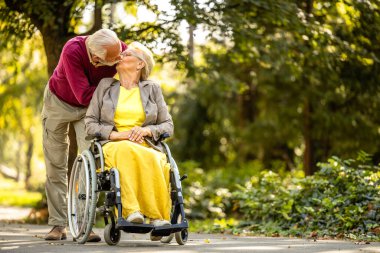 Aşık yaşlı bir çift. Çocuk parkında yaşlı bir adam karısını tekerlekli sandalyede öpüyor..