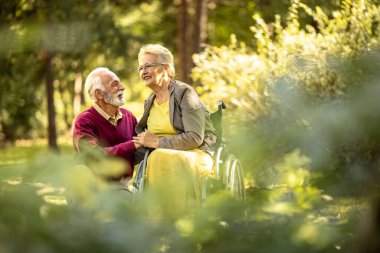 Tekerlekli sandalyedeki yaşlı adam ve karısı çocuk parkında el ele tutuşuyorlar..