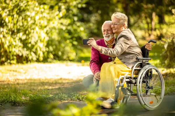 Elderly couple in love. Senior man taking a photo with his wife in wheelchair in nursery home park.