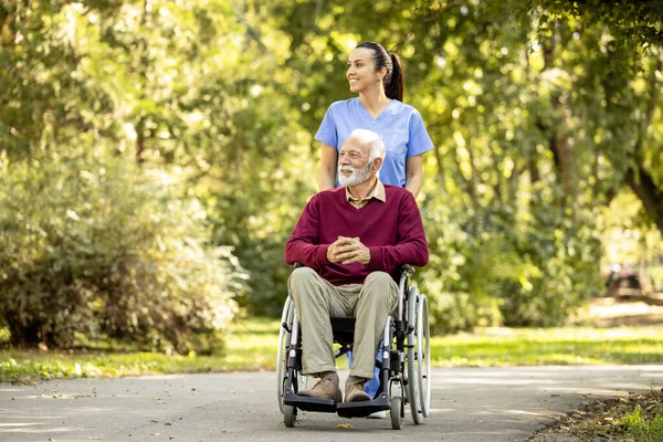 Female nursing assistant and elderly man in wheelchair enjoying autumn in the park.