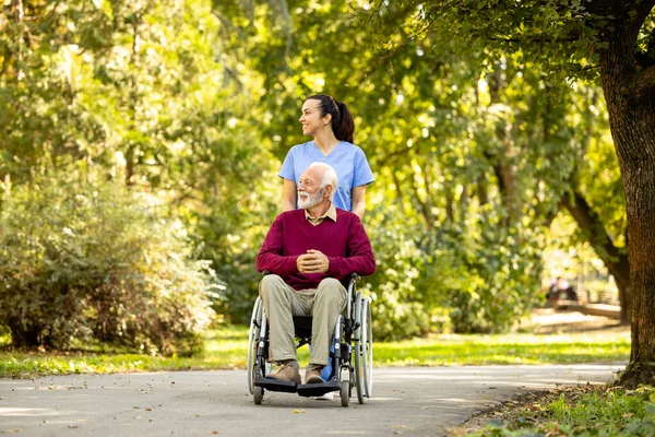 Female nursing assistant and elderly man in wheelchair enjoying autumn in the park.