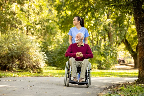 Nursing assistant and elderly man in wheelchair going for a walk in the park.