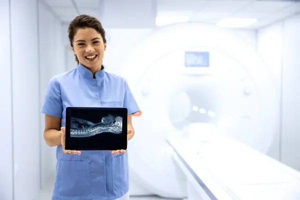 Female doctor radiologist standing in MRI medical examination room and showing X-ray image of patient body.
