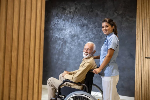 Modern nursing home and female nursing assistant taking care of elderly man in wheelchair.