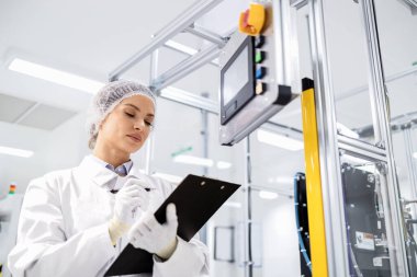 Fabrika mühendisi veya kalite kontrol işçisi üretim makinesinin yanında duruyor ve kontrol listesi tutuyor..