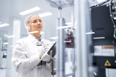 Üretim tesisinde kontrol listesi tutan ve makine operasyonlarını izleyen kadın fabrika işçisi.