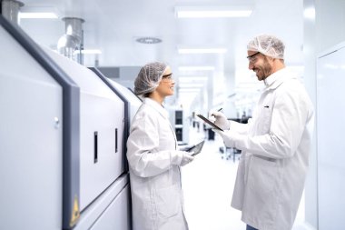 Beyaz önlük ve saç filesi takan mühendisler otomatikleştirilmiş makineleri ve fabrikanın içindeki üretim ekipmanlarını izliyor..