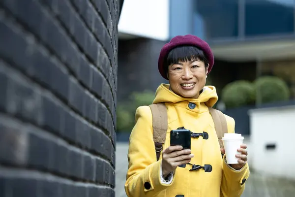 Smiling Japanese woman in coat with coffee cup using mobile phone in city street.