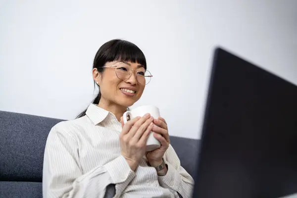 Beautiful Japanese woman having cup of coffee and working on laptop at home.