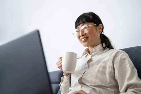 Beautiful Japanese woman having cup of tea and working on laptop at home.