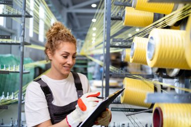 Tekstil fabrikasındaki endüstriyel örgü makinesinin üretimini kontrol eden kadın işçi..