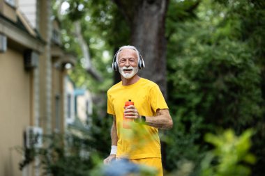 Yaşlı bir adam, gri saçlı, elinde su şişesiyle parkta koşuyor..