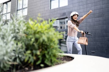 Zarif takım elbiseli gülümseyen kadın şehirde elektrikli scooter sürmenin keyfini çıkarıyor..