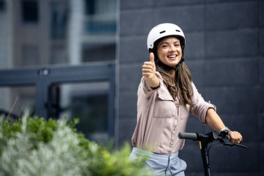 Kasklı gülümseyen bir kadının portresi. Elektrikli scooter 'ın üzerinde duruyor ve başparmağını kaldırıyor..