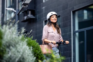 Elektrikli scooter kullanan, kasklı, gülümseyen kadın..