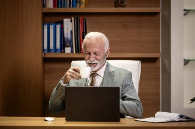 Beyaz sakallı iş adamı lüks ofisinde oturup çalışırken kahve içiyor..
