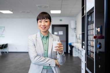 Takım elbiseli bir iş kadını portresi elinde kahve fincanı ve satış makinesinin yanında duruyor..