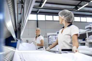 Female laundry workers folding linen and bedsheets after ironing in dry cleaning service. clipart