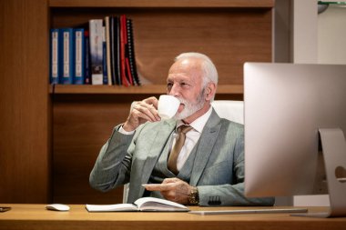 Beyaz sakallı iş adamı lüks ofisinde oturup çalışırken kahve içiyor..