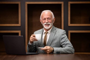 Gülümseyen yakışıklı kıdemli işadamının portresi rahat ofis koltuğunda oturuyor ve kahve içerken dizüstü bilgisayarda çalışıyor..