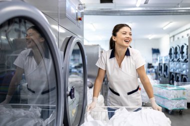 Gülümseyen kadın işçi çamaşır sepetini tutuyor ve kuru temizleme servisinde çamaşır makinesi çalıştırıyor..
