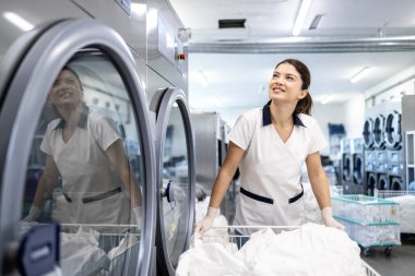 Gülümseyen kadın işçi çamaşır sepetini tutuyor ve kuru temizleme servisinde çamaşır makinesi çalıştırıyor..