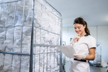 Hastane çamaşırhanesinde çamaşır arabasının yanında duran kontrol listesi olan bir kadın işçi..