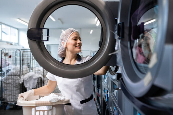 Professional laundry washing and drying service. Female worker standing by washing machine and sorting clothes.