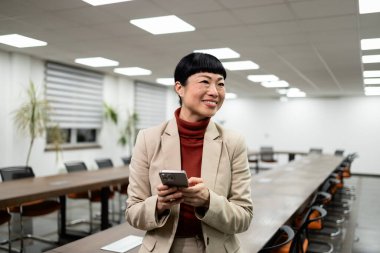 Şık takım elbiseli gülümseyen Japon kadın şirket toplantı odasında telefonunu kullanıyor..