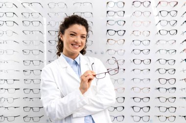 Optik mağazasında gözlük panelinin önünde duran güzel, gülümseyen kadın gözlükçünün portresi.