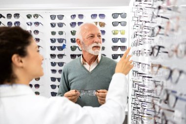 Tecrübeli bayan gözlükçü, optik mağazada yeni gözlük için mükemmel çerçeveyi seçmesine yardım ediyor..