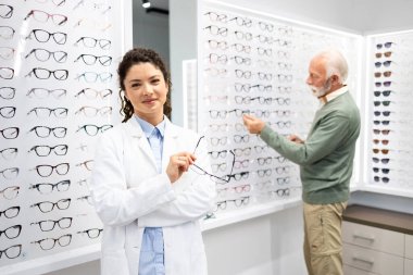Beyaz önlüklü güzel kadın gözlükçünün portresi optik mağazada dikilirken arka planda yaşlı adam gözlük çerçevesini seçiyor..