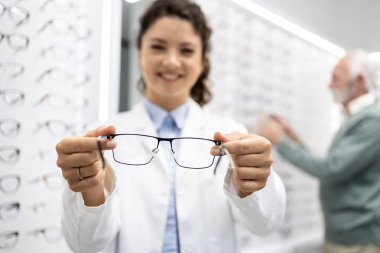 Güzel gülümseyen optometrist, gözlük dükkanında modern gözlük tutarken arka planda yaşlı hasta bir çift gözlük seçiyor..
