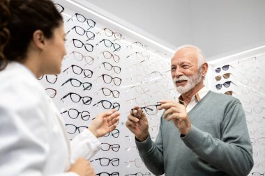Güzel bayan optometrist kıdemli hastaya optik mağazasından uygun bir çift gözlük seçmesini tavsiye ediyor..